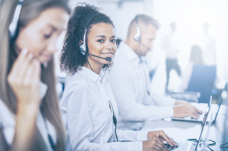 Call center employee using Sharepoint and wearing a headset and surrounded by her team. She is smiling and look at the photographer.