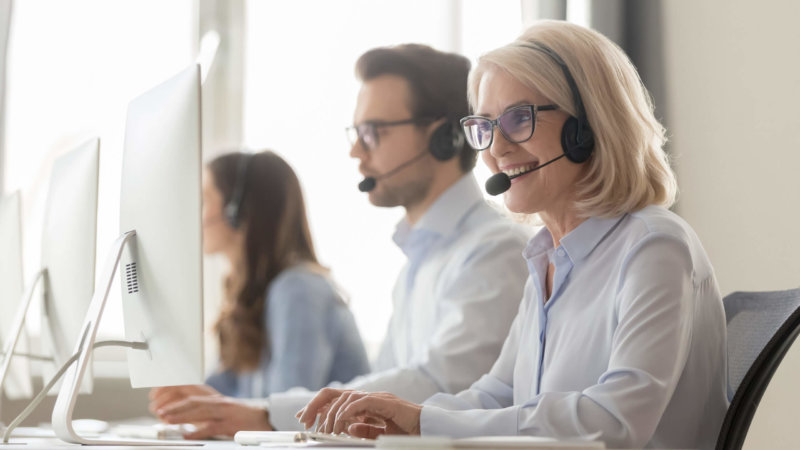 Photo of three contact center employees at work on computers, using headsets, and smiling // contact center software features concept
