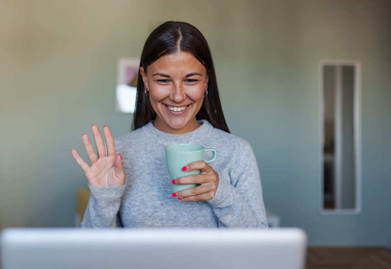Photo of woman sitting in front of a laptop having a web conferencing meeting, she is drinking a cup of coffee and waving at her peers. Tools for remote teams concept.